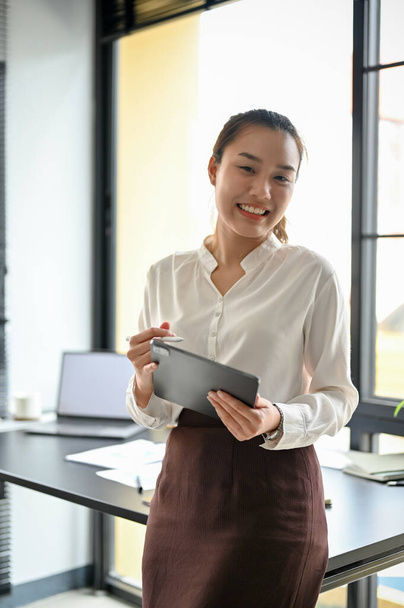 Portrait of a friendly Asian female manager, female boss leaning on her office desk, holding a digital tablet touchpad. - Photo, Image