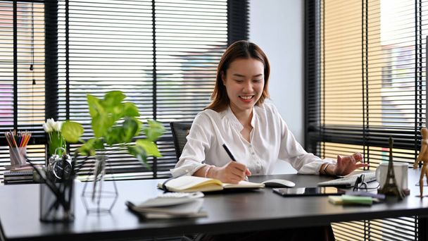 Cheerful Asian female manager sits at her desk in a private office workplace, working and managing her tasks. - Foto, Bild