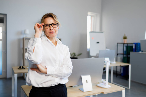 Αυτοπεποίθηση ώριμη επιχειρηματίας με γυαλιά και λευκό πουκάμισο στέκεται στο γραφείο - Φωτογραφία, εικόνα