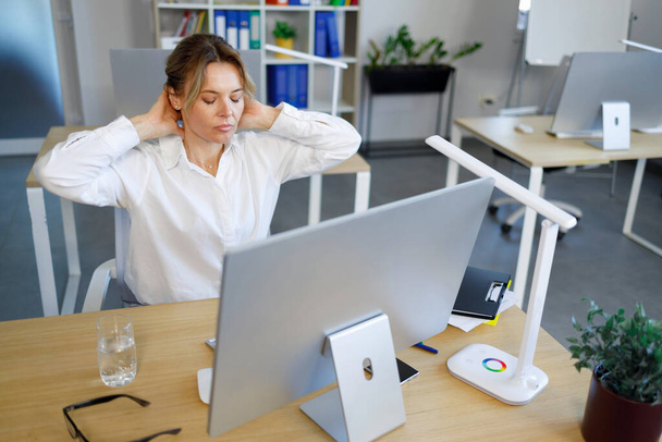 Жіночий офісний працівник розтягується для відпочинку під час роботи за комп'ютером. Ділова жінка масажує шию, сидячи на робочому місці
 - Фото, зображення