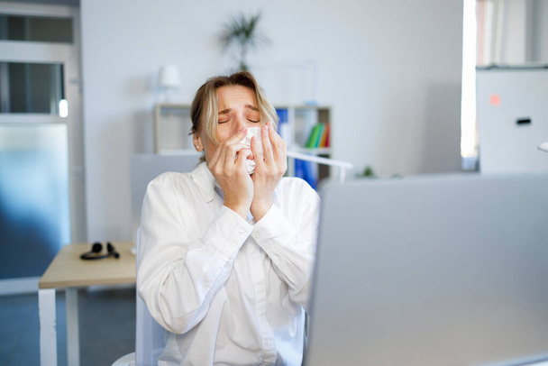Sick businesswoman sneezing in a tissue while working in the office - Fotoğraf, Görsel