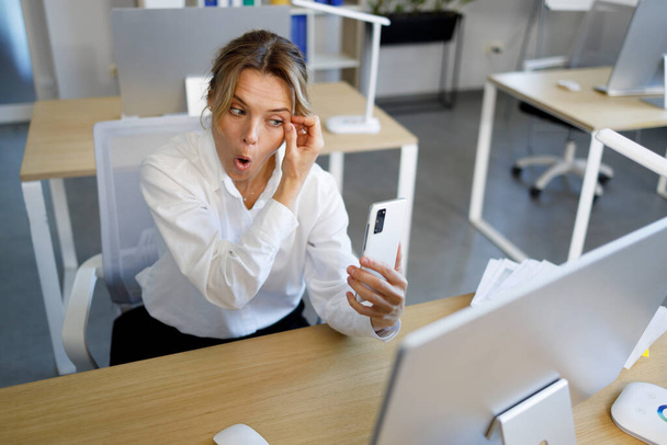 Γραφείο γυναίκα προσαρμογή μάσκαρα μπροστά από τα μάτια της, ενώ κοιτάζοντας κάμερα smartphone - Φωτογραφία, εικόνα