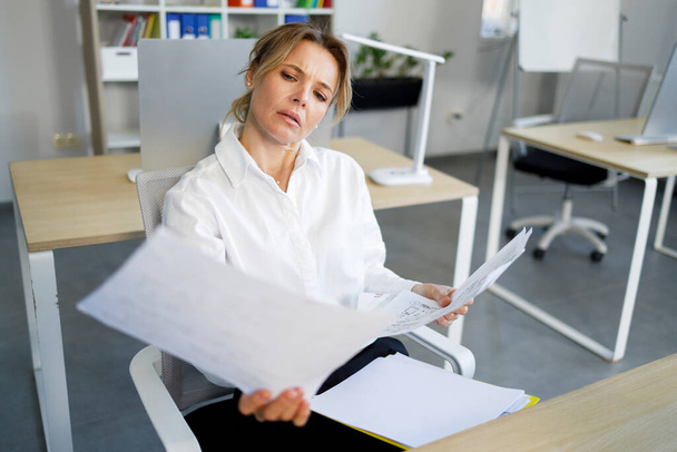 volwassen kantoor vrouw in formele dragen examen documenten terwijl zitten op haar werkplek - Foto, afbeelding