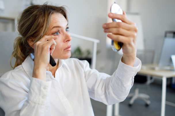 Business woman looks in a cosmetic mirror and touch up her make-up of her eyes  - Fotoğraf, Görsel
