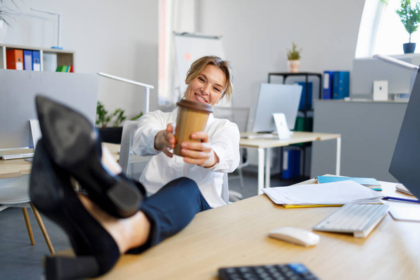 Happy business woman with cup of coffee relaxes putting legs on desk - Foto, Imagen