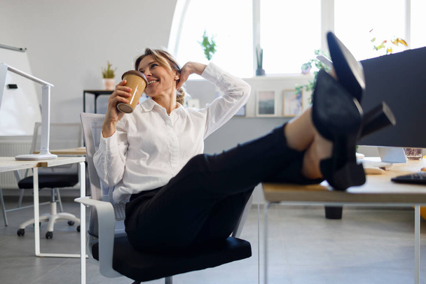 Happy business woman drinking coffee putting legs on the table - Photo, image