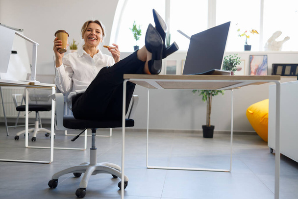 リラックスしたビジネス女性とともにコーヒーとクッキーはテーブルの上に彼女の足を置く  - 写真・画像