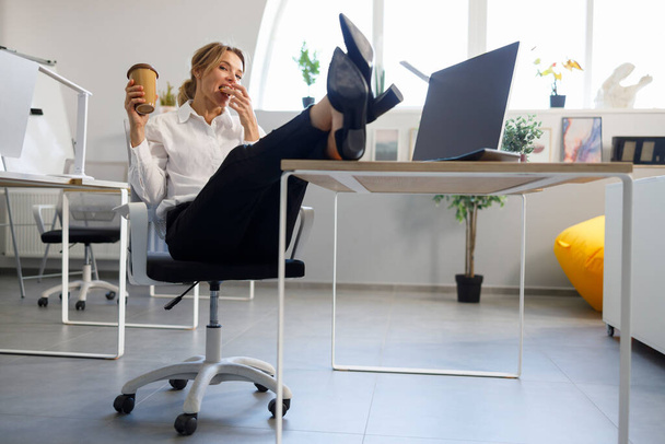 Business woman with a cup of coffee put her legs on the desk and eats cookies - Foto, imagen