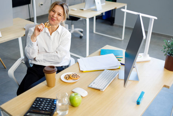 Ενηλίκων γυναίκα υπάλληλος γραφείου τρώει μπισκότα κατά τη διάρκεια του διαλείμματος καφέ - Φωτογραφία, εικόνα