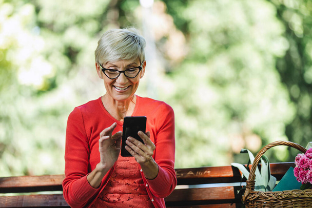 Upea hymyilevä valkoihoinen vanhempi nainen vaaleat lyhyet hiukset istuu penkillä puistossa ja soittaa puhelun älypuhelimella. - Valokuva, kuva