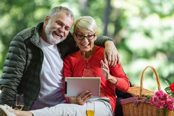Feliz pareja de ancianos haciendo un picnic en el parque usando tableta digital - Foto, imagen