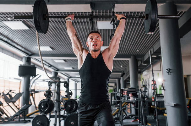 Lifting barbell. Young sportive strong man in black wear have workout day in gym. - Foto, Bild