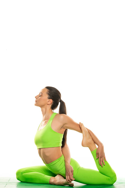 Молодая спортивная привлекательная женщина практикующая йогу, делающая упражнения Русалка, Eka Pada Rajakapotasana поза, разминка, носить спортивную одежду, зеленые брюки и топ, крытый полный рост, белый фон.. - Фото, изображение