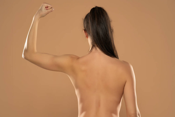 Rückenansicht einer jungen nackten Frau mit Pferdeschwanz, die den Arm auf beigem Hintergrund zeigt. - Foto, Bild