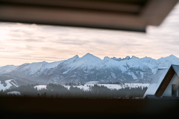 roof window view of sunset in the mountains. view from the attic window of the wooden cottage in mountain valley. Panorama of snowy mountains. - Fotoğraf, Görsel