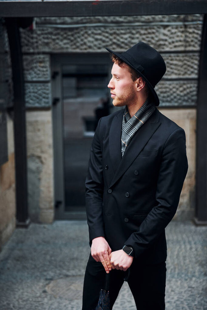 Elegant young man in black hat and clothes with umbrella in hands outdoors in the city. - Foto, immagini