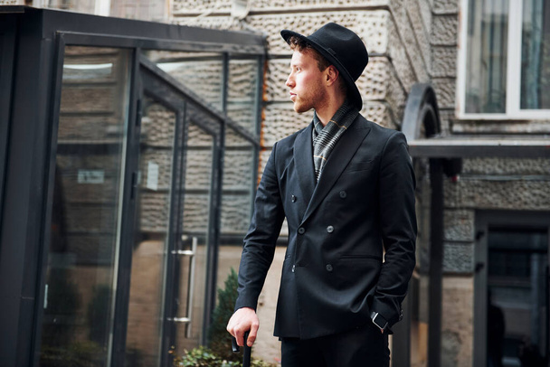 Elegant young man in black hat and clothes with umbrella in hands outdoors in the city. - Valokuva, kuva