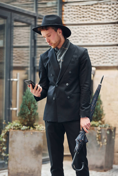 Talking by phone. Elegant young man in black hat and clothes with umbrella in hands outdoors in the city. - Photo, Image