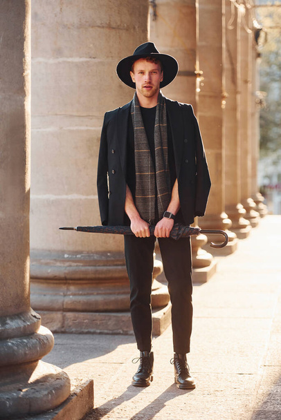 Elegant young man in black hat and clothes with umbrella in hands outdoors in the city. - Photo, image