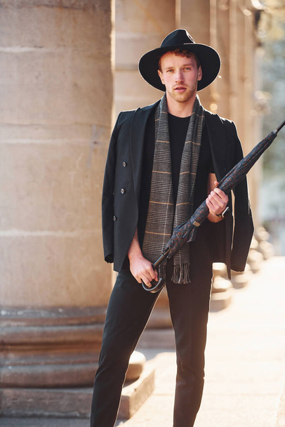 Elegant young man in black hat and clothes with umbrella in hands outdoors in the city. - Photo, Image