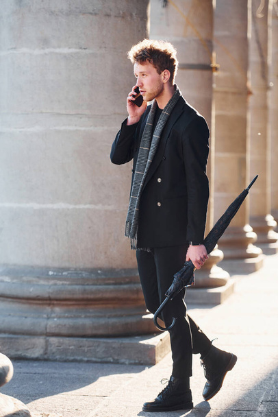 Elegant young man in formal classy clothes standing and talking by the phone outdoors in the city. - Foto, afbeelding