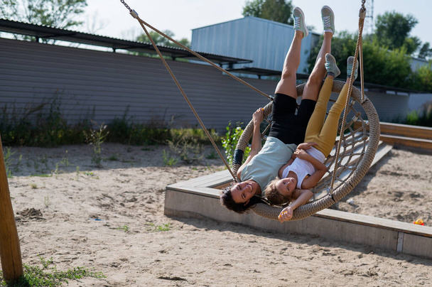 Mom and daughter swing on a round swing. Caucasian woman and little girl have fun on the playground. - Φωτογραφία, εικόνα