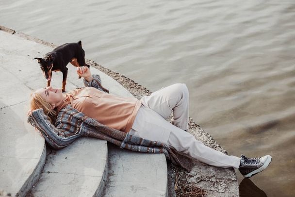 Mladá blondýnka v teplých šatech ležící na břehu řeky s pěkným štěňátkem za chladného slunečného dne, odpočívající a užívající si boční pohled. Venkovní víkendové aktivity, láska a péče o zvířata, vodní zázemí - Fotografie, Obrázek