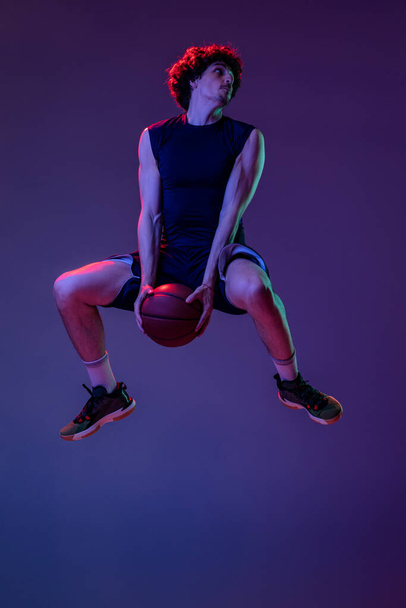 Dynamic portrait of professional basketball player jumping with ball isolated on purple background in neon. Concept of sport, art, creation, competition. - Foto, immagini