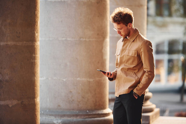 Holding phone in hand. Elegant young man in formal classy clothes outdoors in the city. - Photo, Image