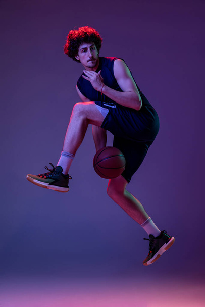 Dynamic portrait of professional basketball player jumping with ball isolated on purple background in neon. Concept of sport, art, creation, competition. - Fotoğraf, Görsel