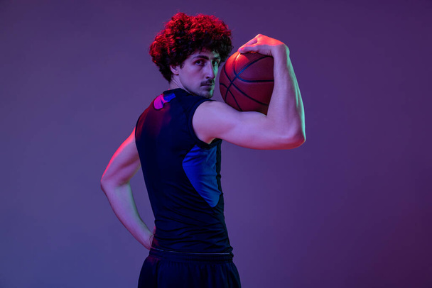 Back view of male muscular basketball player wearing blue sportswear isolated on purple background in neon. Concept of sport, art, creation, competition. - Foto, Bild