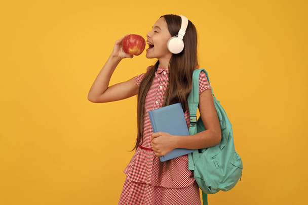 χαρούμενο παιδί του σχολείου με ακουστικά κουβαλάει σακίδιο πλάτης και βιβλίο εργασίας τρώει μήλο για μεσημεριανό, τρώει - Φωτογραφία, εικόνα