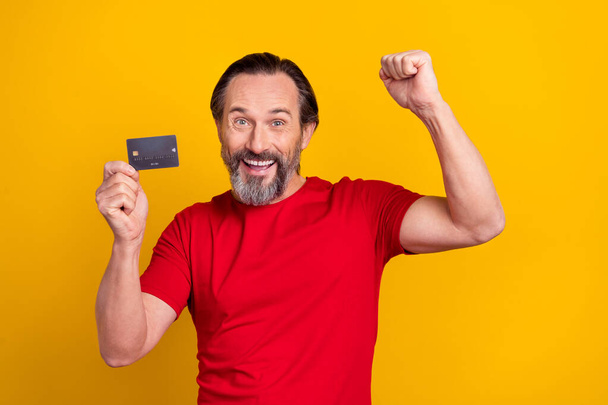Photo of lucky funky retired man wear red t-shirt rising fist bank card isolated yellow color background - Photo, Image