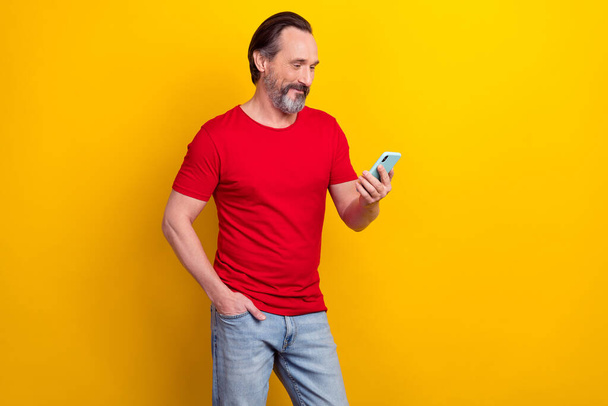 Photo of funny attractive guy pensioner dressed red t-shirt typing modern device isolated yellow color background - Photo, image