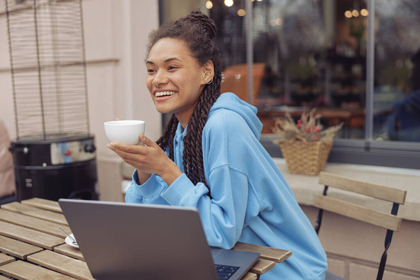 Beautiful young mixed-race cheerful woman with piercing holding coffee, smiling. Cafe terrace. - Photo, image