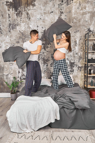 Cheerful pregnant woman cheer handsome guy married couple in love playing with pillows, sheets, bed, enjoy Saturday morning weekend, good excited mood, wear pajama room indoors. - 写真・画像
