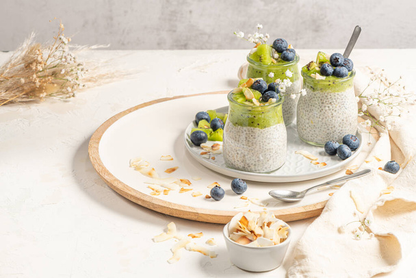 Healthy breakfast chia pudding with kiwi, blueberries and coconut slices, three portions in glass jars on a white table. - Zdjęcie, obraz
