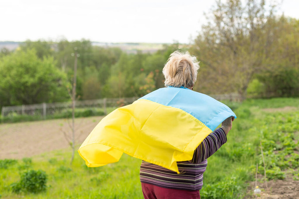 Senior white-haired woman wearing eyeglasses standing outdoors in the country waving the Ukrainian flag looking away feeling the freedom. No war, stop fights, we want peace. - Photo, Image