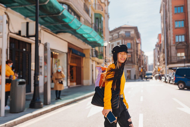 Asian woman, with backpack and mobile phone in hand, doing tourism in a European city. Happy Chinese girl walking down the street and crossing a road. - Zdjęcie, obraz