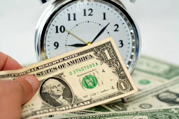 Paying American money banknote currency bills, USA dollars with a blurred alarm clock timer in hours, minutes and seconds scale in the background, paying money for time, time is money concept - Photo, image
