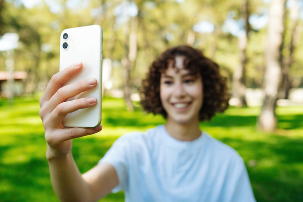 Photo of happy redhead young woman holding phone make selfie or taking video for social media on city park, outdoors. Good mood looks into camera and takes selfie. Selective focus on phone. - Foto, afbeelding