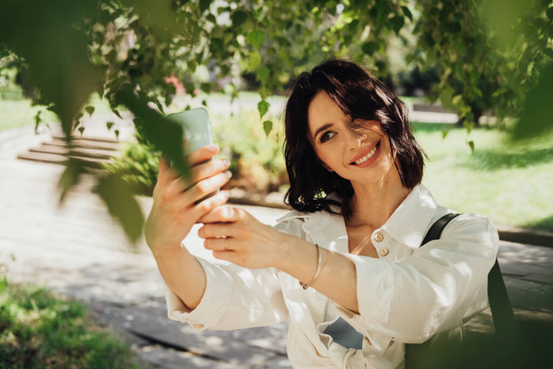 Schöne junge Frau macht Selfie auf Smartphone im Freien unter Bäumen im Park bei sommerlichem Sonnenschein - Foto, Bild