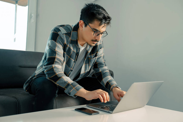 Молодий чоловік в сорочці, сидячи на дивані вдома, працює на ноутбуці онлайн, використовуючи Інтернет, усміхнений, щасливий настрій, фрілансер, вільний час дозвілля, спокійний, сучасний спосіб життя роботи
 - Фото, зображення