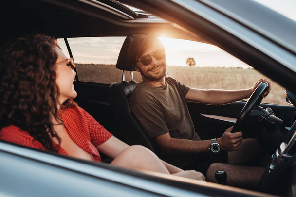 Χαρούμενος άνθρωπος και σγουρά μελαχρινή γυναίκα κάθεται στο αυτοκίνητο, νεαρό ζευγάρι απολαμβάνοντας το οδικό ταξίδι στο ηλιοβασίλεμα - Φωτογραφία, εικόνα