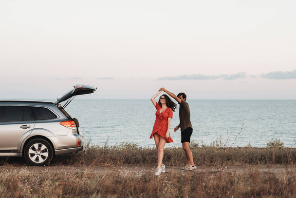 Gençlik Çifti Denizin Arkasında Eğleniyor, Erkek ve Kadın Gün batımında SUV arabalarının yanında Dans Ediyor - Fotoğraf, Görsel