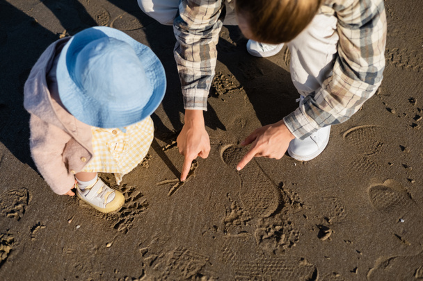 Vista aerea di papà che disegna sulla sabbia vicino al bambino in cappello panama sulla spiaggia  - Foto, immagini