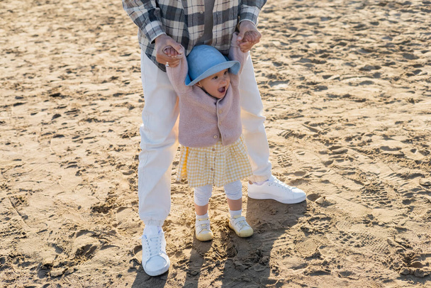 Mann hält fröhliches Baby mit Panamahut am Strand an der Hand  - Foto, Bild