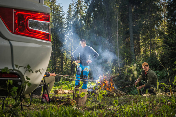 Caucasian Friends in Their 40s Hanging Out Next to Campfire and RV Camper Van in Scenic Woodland Wilderness. Recreational Vehicle Dry Campsite. - Photo, Image