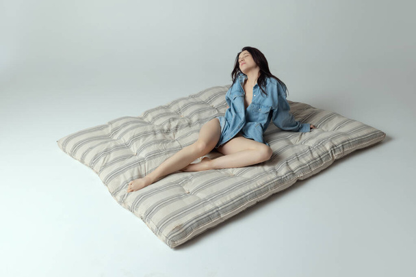 Eine schöne junge Frau sitzt auf einer Matratze, posiert in Unterwäsche und Hemd isoliert vor grauem Studiohintergrund. Natürliche Schönheit - Foto, Bild