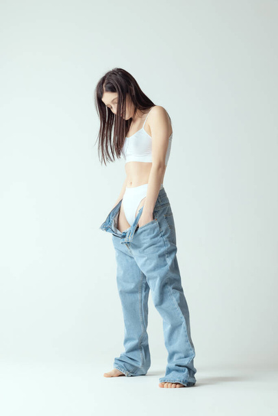 Full-length portrait of young slim woman wearing big jeans, posing isolated over grey studio background. Weight-loss concept - Foto, immagini